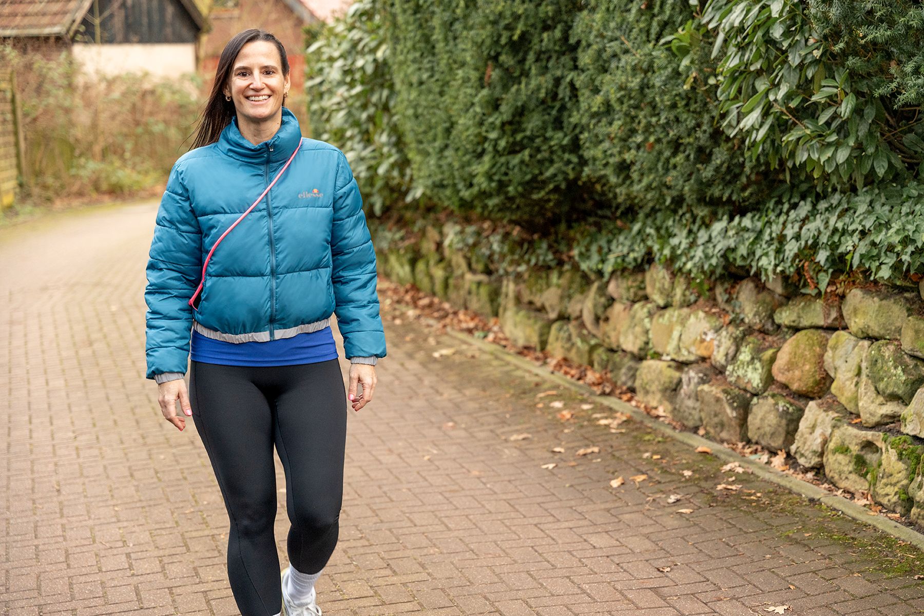 Frau in blauer Jacke und Leggings, die auf einem gepflasterten Weg in einer natürlichen Umgebung spaziert.